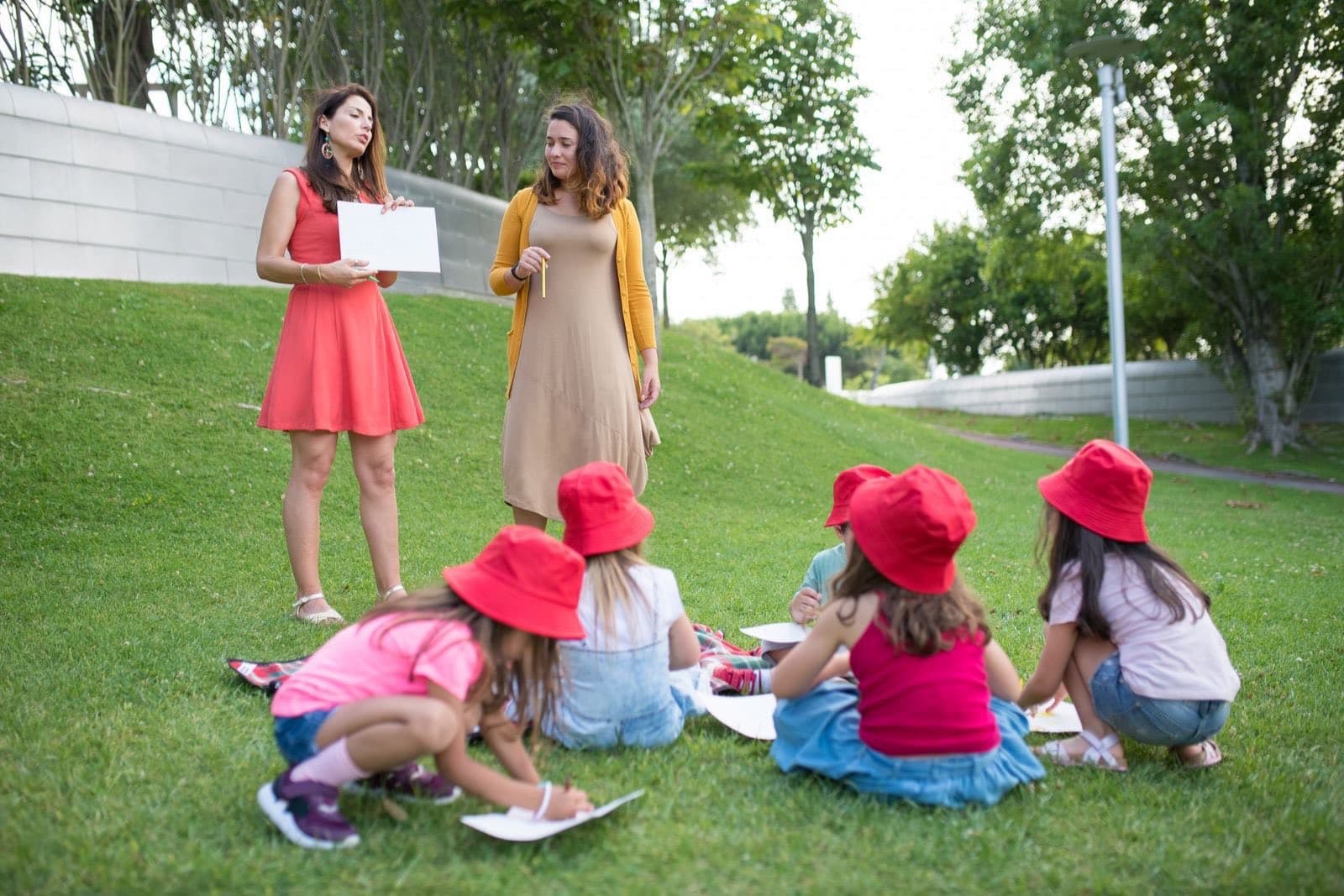 Two adults leading a group of children in red sun hats through a drawing activity on the grass